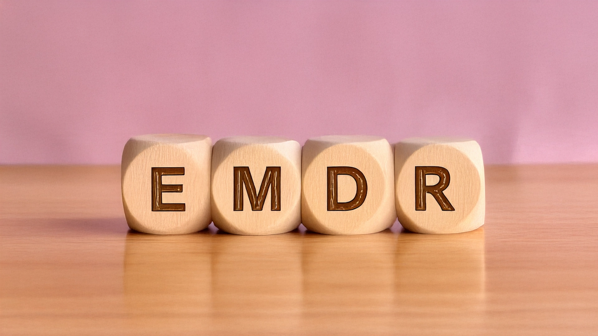 Four dice spelling out the acronym, EMDR on a wooden table with a pink background.